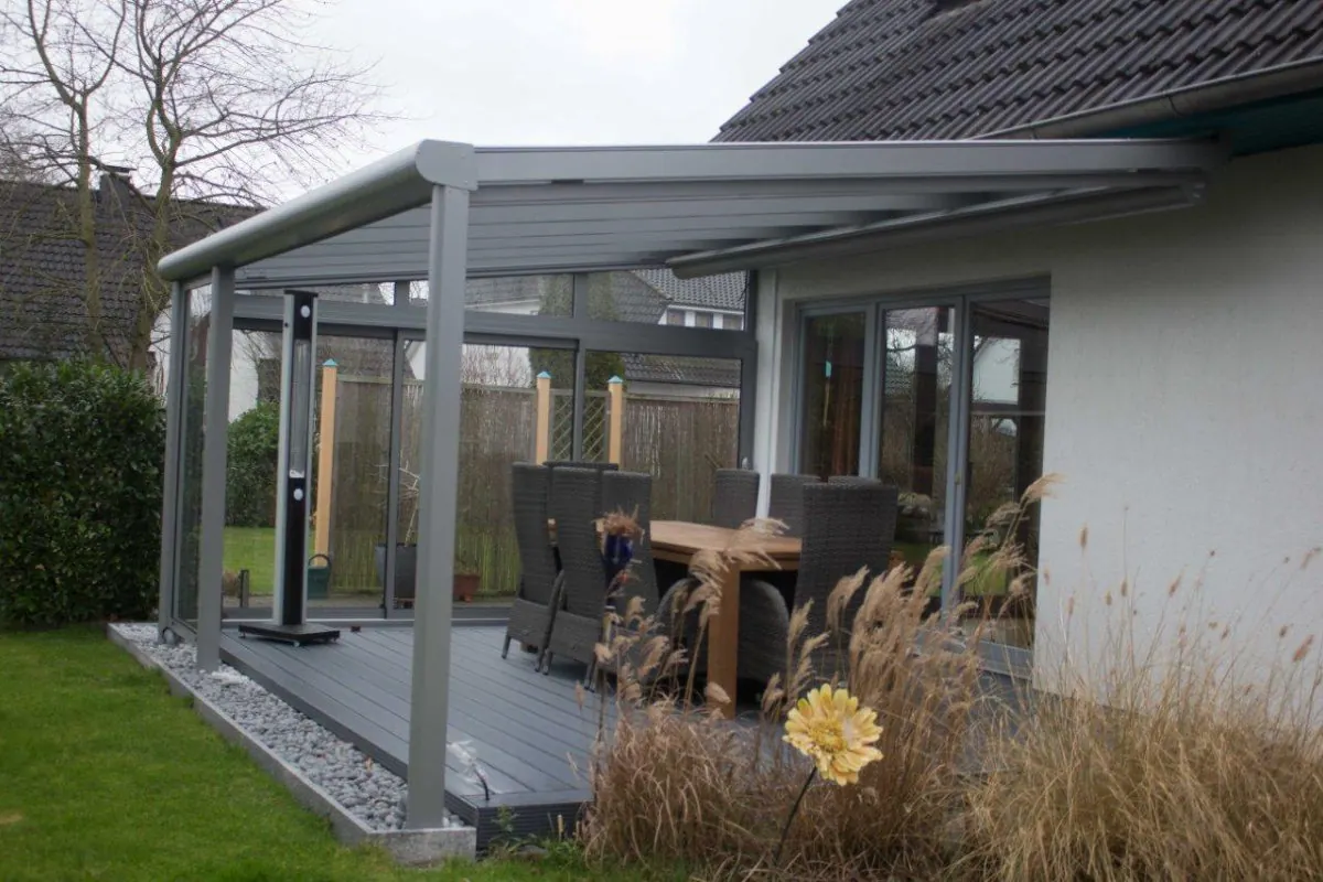 Terrassenüberdachung mit seitlichen Glaselementen und Esstisch vor grauem Wohnhaus