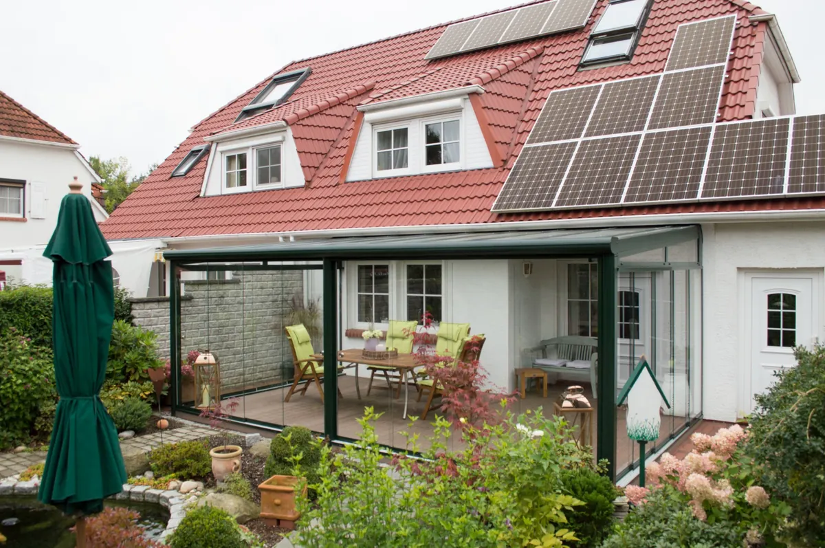 Grüner Sommergarten mit verglaster Terrasse und Sitzbereich vor einem Haus mit rotem Dach