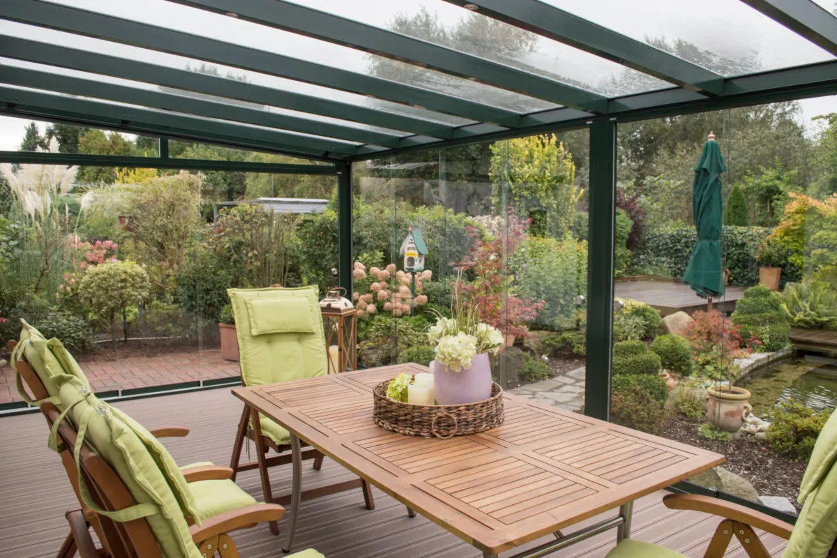Geschützter Sommergarten mit Glaswänden, Tisch und Stühlen sowie Blick in einen bepflanzten Garten